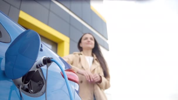 白人女性は電気自動車のトランクの近くに立っており 車両が充電されるのを待っている 電気自動車の充電プラグ — ストック動画