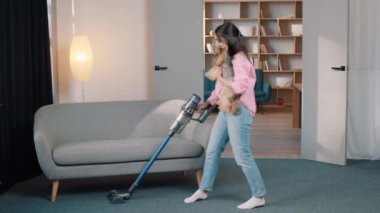 Elinde elektrik süpürgesi olan genç ve çekici bir kız kucağında küçük bir köpekle odayı temizliyor.