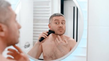 Aynadaki yansımanın arkasındaki erkek aynası. Banyodaki çıplak adam. Sakallı beyaz erkek esmer. Evde tıraş olurken çenesini tıraş etmek için elektrikli tıraş makinesi kullanıyor.