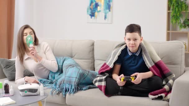 一位年轻妇女在家里用吸入雾化器躺在沙发上 小男孩用操纵杆玩游戏 — 图库视频影像