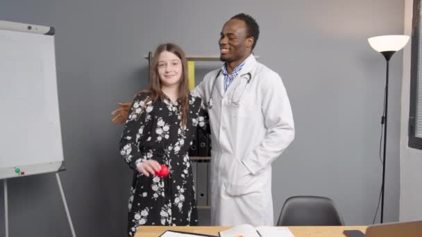 Черный Доктор Обнимает Молодого Пациента Медицинском Кабинете Девушка Показывает Красное — стоковое видео