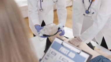 Beyaz laboratuvar önlüklü ve mavi eldivenli bir grup doktor klinikteki mankenlere ilk yardım yapıyor.