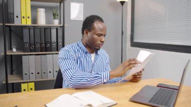 Belgeleri olan genç bir Afrikalı çalışan, mektup alıyor, yasal kağıtları okuyor, proje raporlarını inceliyor, dizüstü bilgisayarında oturuyor.