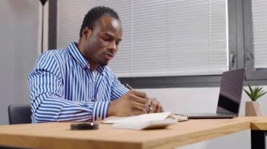 Ciddi genç Afrikalı işadamı belgelerle çalışıyor, bilgisayarına bakıyor ve ofisteki bir deftere yazıyor.