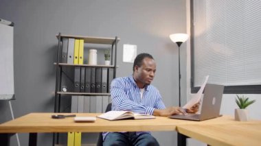 Belgeleri olan genç bir Afrikalı çalışan, mektup alıyor, yasal kağıtları okuyor, proje raporlarını inceliyor, dizüstü bilgisayarında oturuyor.