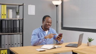 Ofiste akıllı telefon kullanan siyah Afrikalı Amerikalı. Mutlu Adam gülümsüyor ve iş arkadaşları ve müşterileriyle konuşuyor.