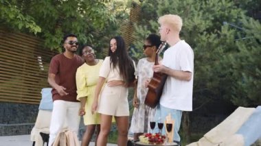 Bir grup arkadaş, sakin bir açık alanda rahatlıyor, kahkahalarını paylaşıyor, dans ediyor.