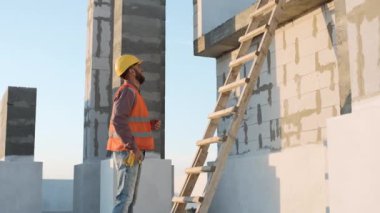 Öğleden sonra inşaatın ilerleyişini merdiven ve güvenlik teçhizatıyla değerlendiren bir işçi.