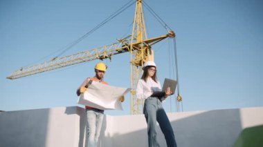 İki inşaat işçisi planları inceliyor ve gündüz vakti bir inşaat projesinde teknolojiyi kullanıyorlar.