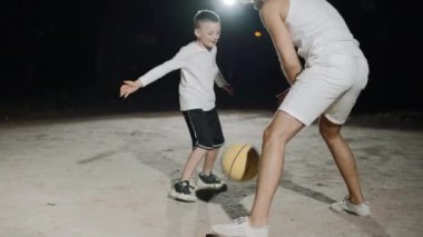 Baba ve oğlu arka bahçede gece lambaları altında basketbol oynuyorlar.