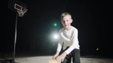 Gece vakti, açık hava ışıkları altında ailesiyle basketbol becerilerini yavaş çekimde uygulayan bir çocuk.