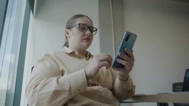 Ofis ortamında bir masada ağır çekimde otururken akıllı telefon kullanan iş elbiseli bir kadın.
