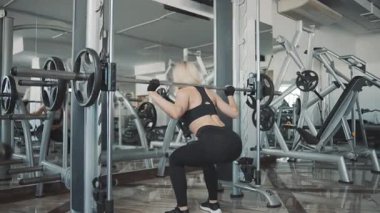 Güçlü kız modern spor salonunda çeşitli spor ekipmanlarıyla ağır çekimde çömelme hareketleri yapıyor.
