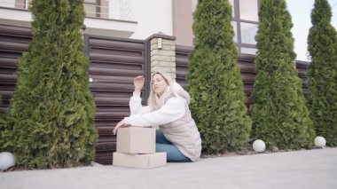 Teslimatçı kadın bir evin dışında kutuların yanında oturup telefonla konuşuyor.