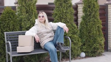 Teslimatçı kadın güneşli bir günde konutta kutularla bankta bekliyor.