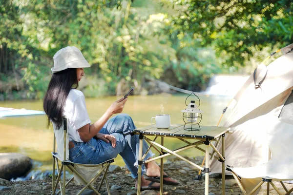 Camping outdoor. Asian women camping leisure and destination travel near waterfall. People hand holding smartphone and drinking coffee on the tents in morning. Tourism relax and chill in summer holiday