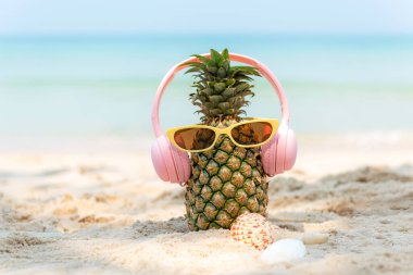 Partide yaz mevsimi. Güneş gözlüklü hippi Pineapple Fashion ve kumsalda güzel mavi gökyüzü arka planında güneş kremi ve sandaletle müzik dinlemek. Moda Yaz Tatili Konsepti