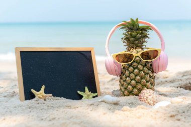 Partide yaz mevsimi. Güneş gözlüklü hippi Pineapple Fashion ve kumsalda karatahta güzel mavi gökyüzü arka planında müzik dinlemek. Moda Yaz Tatili Konsepti