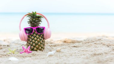 Partide yaz mevsimi. Güneş gözlüklü hippi Pineapple Fashion ve kumsalda güzel mavi gökyüzü arka planında güneş kremi ve sandaletle müzik dinlemek. Moda Yaz Tatili Konsepti, afiş için kopyalama alanı