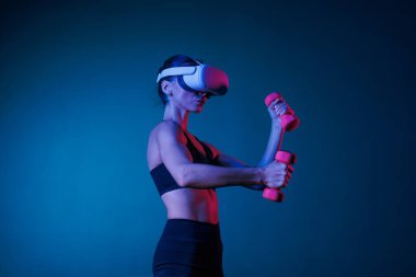Atletik kız sanal gerçeklik gözlüğü takıyor siyah elbiseli koyu renkli dambılı, online spor eğitimi alıyor.