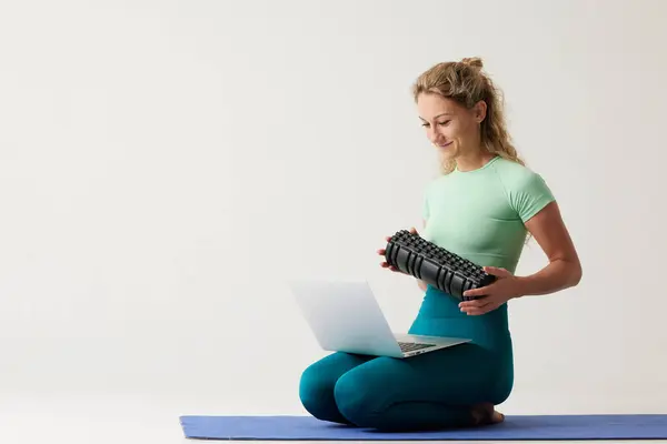 girl holding a massage roller while sitting on the floor and near a laptop, massage roller for fitness and yoga, home online workouts