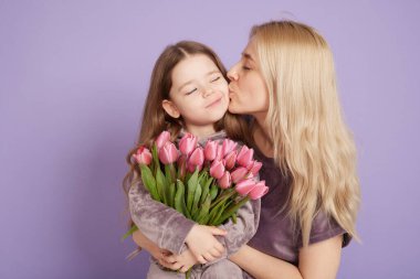 Annesiyle küçük bir kız ve bir buket çiçek mor bir arka planda sarılıyorlar, aile tatili ve birliktelik..