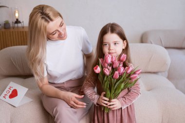 Anneler Günün kutlu olsun! Çocuğun kızı annesini tebrik eder ve ona çiçek laleleri ve bir kart verir. Anne ve kız gülümseyip sarılıyor. Aile tatili ve birliktelik.
