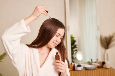 Genç bir kadın pipetinden bir damla yağı saçına sürüyor, yakın plan. Vitaminler, tedavi için keratin, saç güçlendirici ve büyütme. Kepeklerle ilgili sorunlar, saç dökülmesi. Saç bakımı konsepti