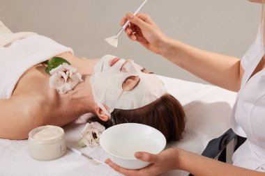 Beautician smoothes sheet mask on woman face for rehydrate face skin, anti wrinkles cosmetic procedure in beauty spa salon. Cosmetologist applying moisturizing sheet mask on female face clipart