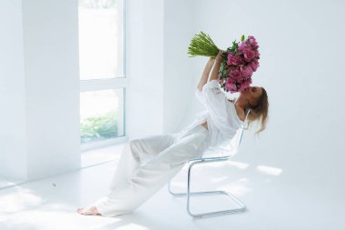 Kocaman bir buket şakayık elinde koca bir demet pembe şakayık çiçekle beyaz arka planda yüzünü ve vücudunu kaplayan bir kadın.