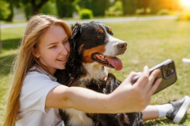 Bir kadın, mutlu ve samimi bir jest yapan sevgili köpeğiyle çimlerin üzerinde selfie çekerek bir anı neşeyle yakalıyor.