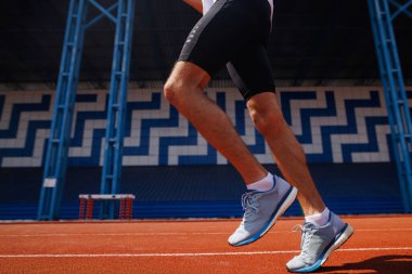 Siyah şortlu ve mavi ayakkabılı bir adam atletizm ve kararlılığını sergileyerek kırmızı bir atletizm pistinde koşuyor.