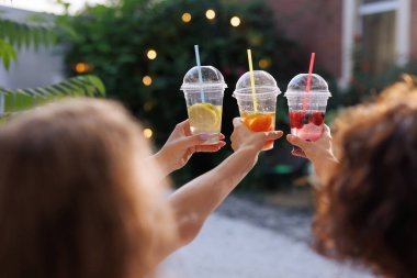 Üç el açık fincanlarda renkli içecekler tutuyor. Pipetle, arka planda hafif ışıklarla bulanık bir şekilde duruyor..