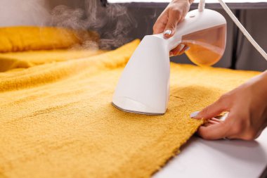 Elle tutulan bir buharlı giysi kullanarak masanın üzerinde pembe bir kumaş buharlaştıran biri. Buhar görünür durumda. Kumaşın yenilendiğini ve kırışıklıkların yok olduğunu gösteriyor..