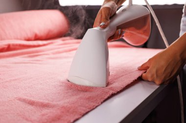 Elle tutulan bir buharlı giysi kullanarak masanın üzerinde pembe bir kumaş buharlaştıran biri. Buhar görünür durumda. Kumaşın yenilendiğini ve kırışıklıkların yok olduğunu gösteriyor..