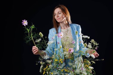 Açık mavi ceketli bir kadın bir buket kır çiçeğinin ortasında duruyor, bir elinde çiçeğe bakıp duruyor. Arka plan koyu renkli, çiçeklerin canlı renklerini ve kıyafetlerini vurguluyor..