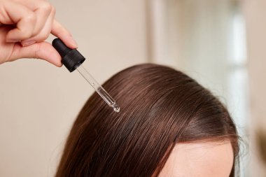 Genç bir kadın pipetinden bir damla yağı saçına sürüyor, yakın plan. Vitaminler, tedavi için keratin, saç güçlendirici ve büyütme. Kepeklerle ilgili sorunlar, saç dökülmesi. Saç bakımı konsepti