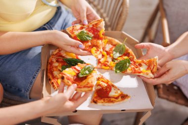 Bir grup arkadaş pizzayı açık havada paylaşıyor, elleri taze fesleğenle süslenmiş dilimlere uzanıyor, pizza kutusu ve mezeler arka planda oturuyor..