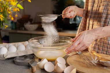Un eleme ve hamuru şeffaf bir kâse içinde mutfağın arka planına karşı hazırlayan kadın ellerine yakın plan. Kutudaki yumurtalar ve hamur yapmak için bir araç masanın üzerinde.