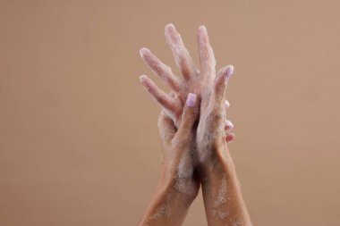 Sabunla sabunlanmış iki elin yakın çekimi, el yıkamayı gösteriyor. Eller, sabun baloncuklarını nötr kahverengi bir arka plana karşı vurguluyor..