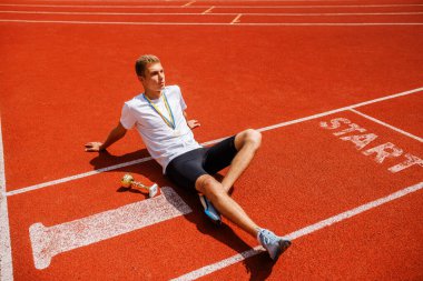 Genç bir erkek atlet koşu pistinde oturuyor, gülümsüyor ve rahatlıyor. Beyaz bir tişört ve siyah bir şort giyiyor, boynunda bir madalya ve yanında da bir kupa var.