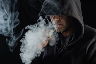 Kapüşonlu adam dumanla çevrili buhar üflüyor. Koyu arkaplan, Elektronik sigara kavramı