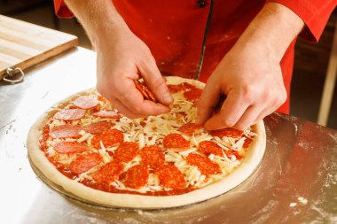 Kırmızı gömlekli biri pepperonili pizza hazırlıyor. Peynir ve domates sosunun üstüne pepperonili dilimler koyuyor..