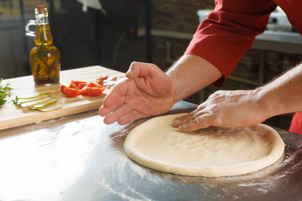 A person shaping pizza dough on a floured surface in a kitchen, with ingredients like tomatoes and pickles in the background.