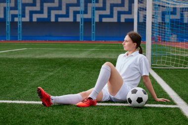 Kadın bir futbolcu futbol sahasının çimlerinde oturuyor, beyaz tişört ve şort giyiyor, kırmızı çizmeleri var. Bir futbol maçından sonra mola
