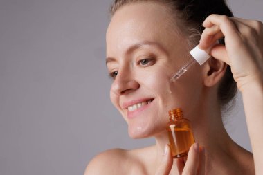 Gülümseyen mutlu kadın yüzüne serum sürüyor, gözleri zevkle kapanıyor. Kızıl saçlı kız deri bakım ürünleriyle dolu bir damlacığı tutuyor sağlıklı ve parlak ten rengi için, mavi arka plan