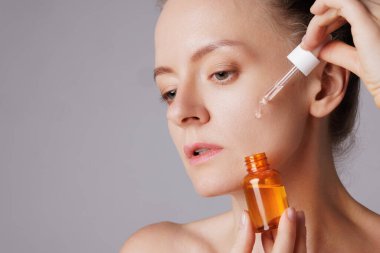 Gülümseyen mutlu kadın yüzüne serum sürüyor, gözleri zevkle kapanıyor. Kızıl saçlı kız cilt bakım ürünleriyle dolu bir damlacığı tutuyor sağlıklı ve parlayan ten rengi için.