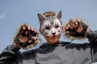 Kedi maskesi ve kürklü eldiven takan neşeli bir çocuk, açık mavi gökyüzüne karşı neşeli bir ifadeyle poz veriyor..