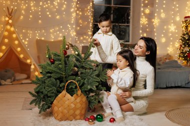 Bir anne ve iki çocuğu küçük bir Noel ağacını peri ışıklarıyla süslenmiş rahat, şenlikli bir odada süslüyorlar. Çocuklar süs eşyalarına neşeyle uzanırken, anne onları gülümseyerek izliyor. Atmosfer sıcak ve neşeli, mükemmel