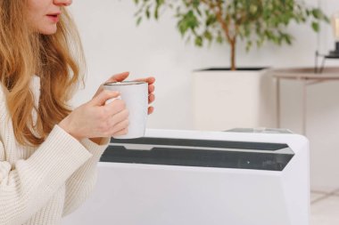 Sıcak ekose ve çay fincanı olan genç bir kadın yatak odasında elektrikli konteyner ısıtıcısının yanında oturuyor, ev ısıtma konsepti, düşük sıcaklık